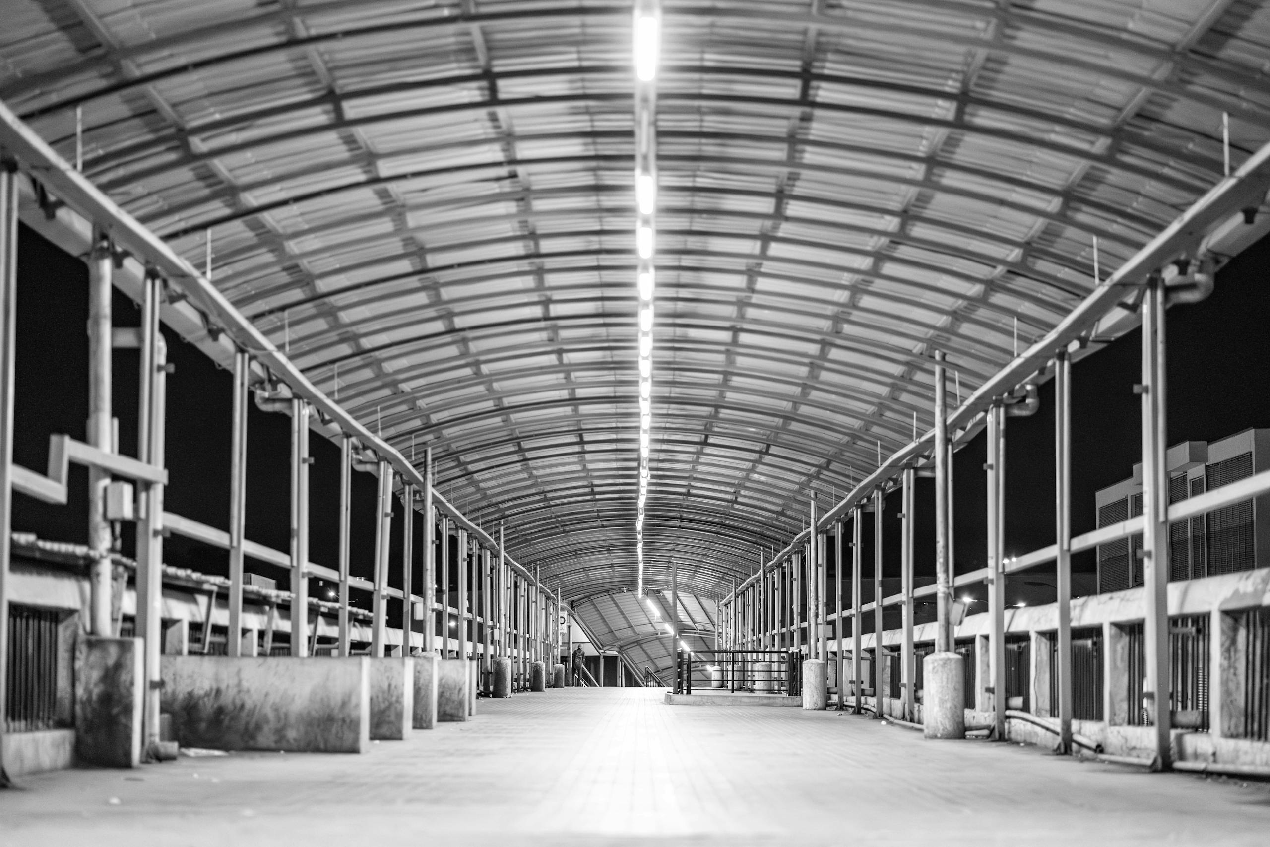 Monochrome image of an empty industrial walkway under a metal roof at night.